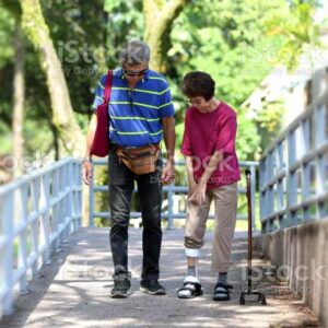 A senior woman is walking at the park with her retiree husband. She had stroke two years ago and currently walking with foot brace as part of rehabilitation exercise. Her husband had eye surgery due to diabetic retinopathy and also stroke survivor.